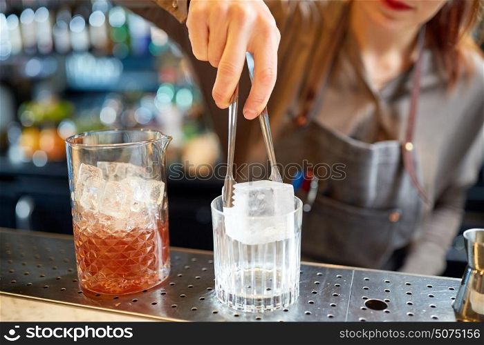 alcohol drinks, people and luxury concept - woman bartender with tongs adding ice cube into glass and preparing cocktail at bar counter. bartender adding ice cube into glass at bar