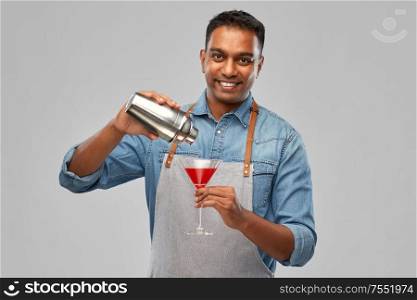 alcohol drinks, people and profession concept - indian barman in apron pouring cocktail from shaker to glass over grey background. indian barman with glass of cocktail and shaker