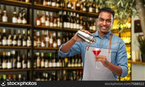 alcohol drinks, people and profession concept - indian barman in apron pouring cocktail from shaker to glass over bar background. indian barman with glass of cocktail and shaker