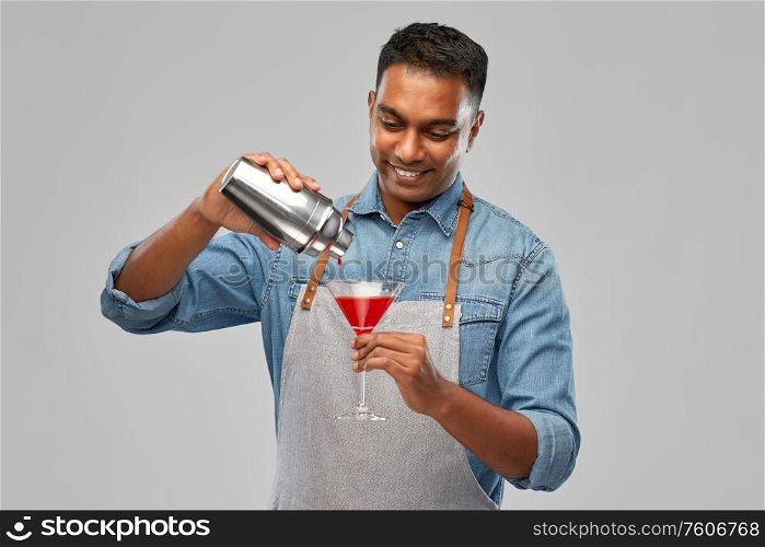 alcohol drinks, people and profession concept - indian barman in apron pouring cocktail from shaker to glass over grey background. indian barman with glass of cocktail and shaker
