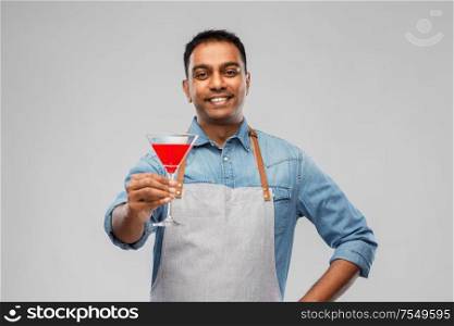 alcohol drinks, people and profession concept - indian barman in apron with glass of cocktail over grey background. indian barman in apron with glass of cocktail