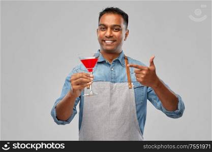 alcohol drinks, people and profession concept - indian barman in apron with glass of cocktail over grey background. indian barman in apron with glass of cocktail