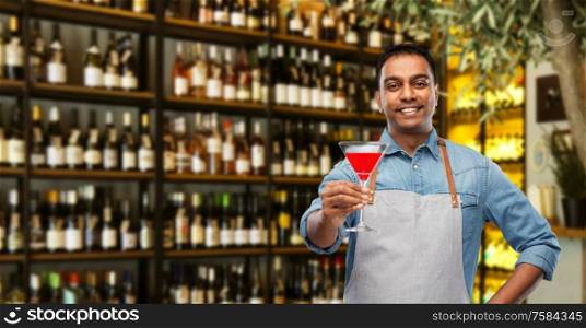alcohol drinks, people and profession concept - indian barman in apron with glass of cocktail over wine bar background. indian barman with glass of cocktail at bar