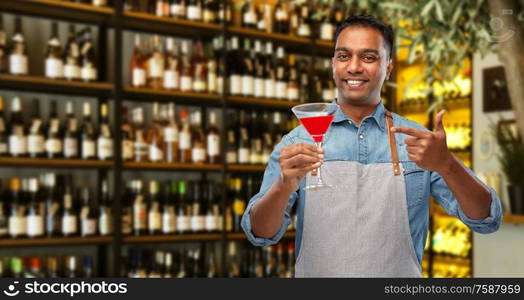 alcohol drinks, people and profession concept - indian barman in apron with glass of cocktail over bar background. indian barman with glass of cocktail at bar