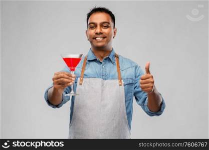 alcohol drinks, people and profession concept - indian barman in apron with glass of cocktail showing thumbs up over grey background. indian barman in apron with glass of cocktail