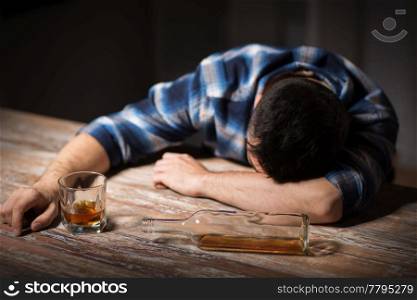 alcoholism, alcohol addiction and people concept - male alcoholic with glass of whiskey and bottle lying or sleeping on table at night. drunk man with glass of alcohol on table at night