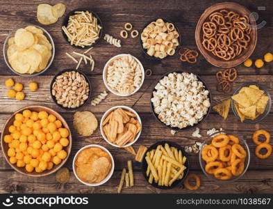 All classic potato snacks with peanuts, popcorn and onion rings and salted pretzels in bowl plates on wood. Twirls with sticks and potato chips and crisps with nachos and cheese balls. Top view