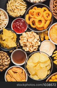 All classic potato snacks with peanuts, popcorn and onion rings and salted pretzels in bowl plates on black table. Twirls with sticks and potato chips and crisps with nachos and cheese balls.