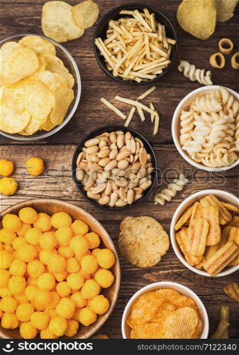 All classic potato snacks with peanuts, popcorn and onion rings and salted pretzels in bowl plates on wood. Twirls with sticks and potato chips and crisps with nachos and cheese balls. Top view