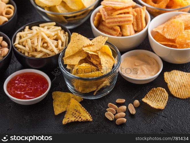 All classic potato snacks with peanuts, popcorn and onion rings and salted pretzels in bowl plates on black table. Twirls with sticks and potato chips and crisps with nachos and cheese balls.
