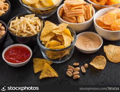 All classic potato snacks with peanuts, popcorn and onion rings and salted pretzels in bowl plates on black table. Twirls with sticks and potato chips and crisps with nachos and cheese balls.