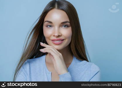 Alluring carefree European female model touches jawline, smiles toothily, has healthy perfect skin, minimal makeup, dreamy gaze, captured with something interesting, isolated over blue background