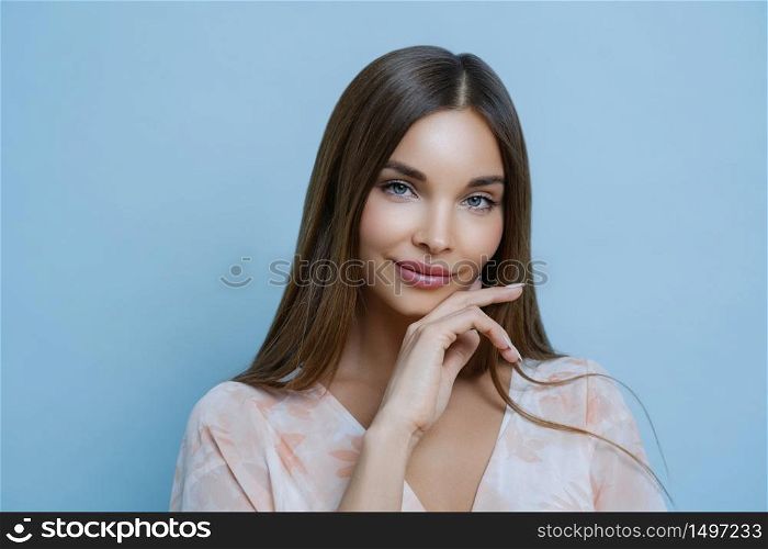 Alluring pleasant looking woman with long dark straight hair touches jawline, looks forward to special event, takes part in profesional photoshoot, has healthy clean skin, poses over blue background