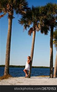 Alluring, sexy woman in feminine dress, standing near ocean leaning on palm trees