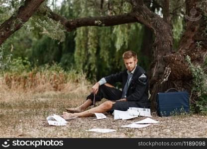 Alone businessman in torn suit sitting on the ground on lost island. Business risk, collapse or bankruptcy concept