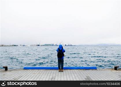 Alone man and calm peaceful sea, Man wear blue hoodie jacket standing at edge of sea port on cloudy foggy winter season day - Explore and travel concept