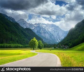 Alone trees and road in alpine mountains, green meadows at sunset in summer. View of country road. Colorful landscape with road, rocks, field, grass, blue sky, sunbeam, clouds. Logar valley, Slovenia
