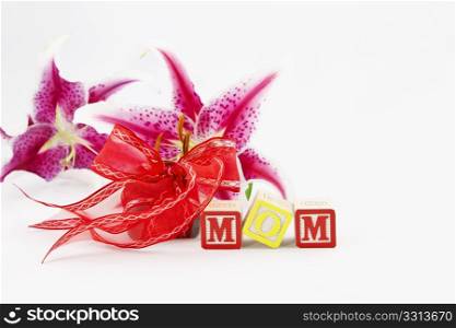 Alphabet blocks spelling out MOM are in front of a red ribbon wrapped gift and two, single lily blooms, all placed on a white background.