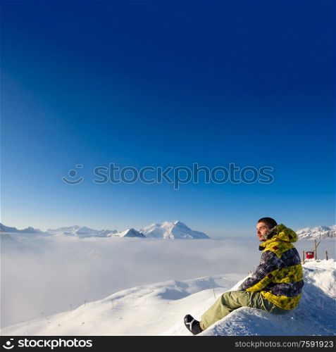 Alpine winter mountain landscape with man sitting above low clouds. French Alps covered with snow in sunny day. Val-d&rsquo;Isere, France