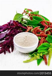 Amaranth flour in a bowl, brown, green and purple flowers of a plant on wooden board background