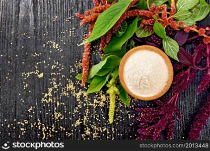 Amaranth flour in a bowl, seeds in a spoon and oil in decanter, brown, green and purple flowers of plant on wooden board background from above