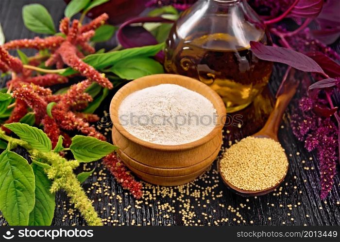 Amaranth flour in a bowl, seeds in a spoon and oil in decanter, brown, green and purple flowers of plant on dark wooden board background