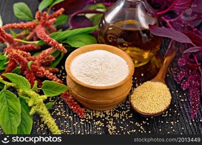 Amaranth flour in a bowl, seeds in a spoon and oil in decanter, brown, green and purple flowers of plant on dark wooden board background