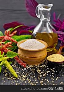 Amaranth flour in a bowl, seeds in a spoon and oil in decanter, leaves and brown, green, purple flowers of plant on wooden board background