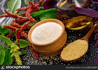 Amaranth flour in a bowl, seeds in a spoon and oil in decanter, leaves and brown, green, purple flowers of plant on dark wooden board background