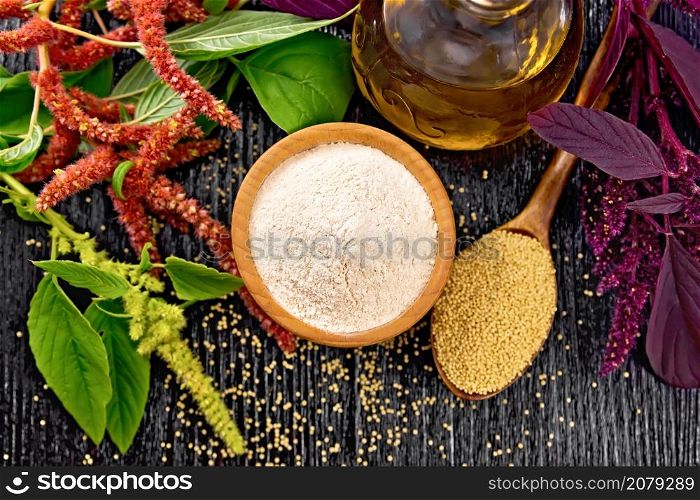 Amaranth flour in a bowl, seeds in a spoon and oil in decanter, brown, green and purple flowers of plant on black wooden board background from above