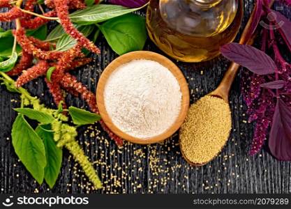 Amaranth flour in a bowl, seeds in a spoon and oil in decanter, brown, green and purple flowers of plant on black wooden board background from above