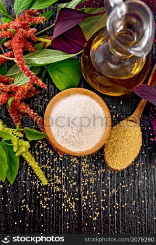 Amaranth flour in a bowl, seeds in a spoon and oil in decanter, brown, green and purple flowers of plant on dark wooden board background from above