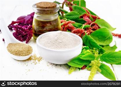 Amaranth flour in a bowl, seeds in a spoon and oil in a glass jar, brown, green and purple flowers of a plant on wooden board background