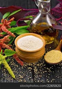 Amaranth flour in a bowl, seeds in a spoon and oil in decanter, brown, green and purple flowers of plant on wooden board background