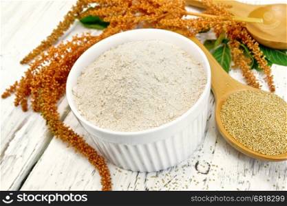 Amaranth flour in white bowl, spoons with grain, brown flower with green leaves on the background light wooden boards