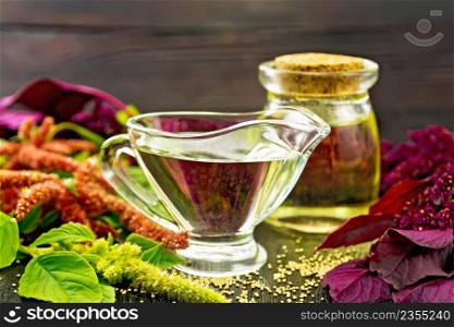 Amaranth oil in a glass gravy boat and jar, brown, green and purple flowers and leaves of the plant on a dark wooden board background