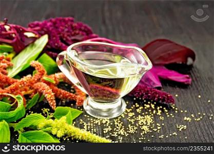 Amaranth oil in a glass gravy boat, brown, green and purple flowers and leaves of the plant on a dark wooden board background