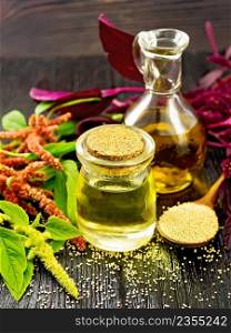 Amaranth oil in a glass jar and decanter, amaranth seeds, brown, green and purple flowers and plant leaves on dark wooden board background