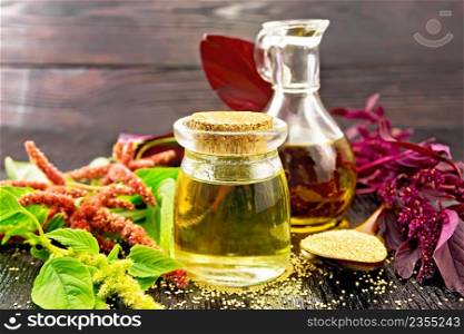Amaranth oil in a glass jar and decanter, amaranth seeds in a spoon, brown, green and purple flowers and plant leaves on a wooden board background