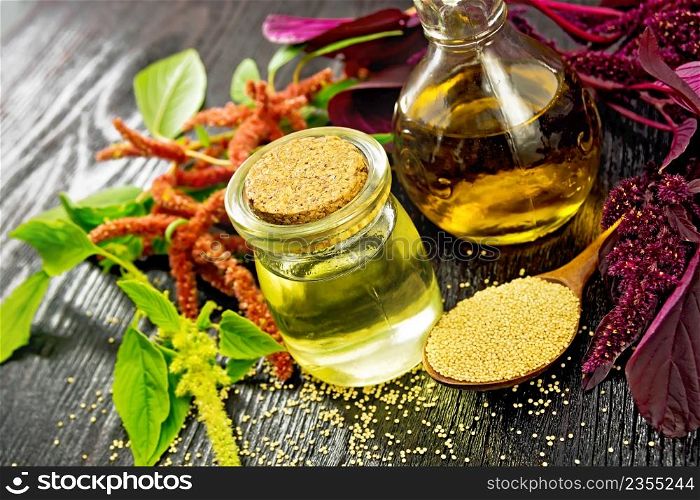 Amaranth oil in a glass jar and decanter, amaranth seeds in a spoon, brown, green and purple flowers and plant leaves on black wooden board background