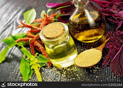 Amaranth oil in a glass jar and decanter, amaranth seeds in a spoon, brown, green and purple flowers and plant leaves on black wooden board background