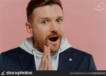 Amazed bearded man in sweatshirt and black jacket, mouth open, palms pressed together, disbelief, isolated on pink background