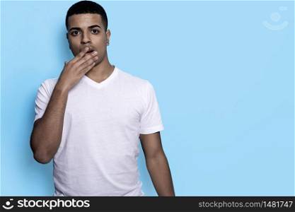 Amazed black young male covering his mouth with palm against blue wall