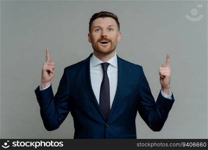 Amazed business leader in suit pointing up, excited with open mouth against grey background, innovative idea
