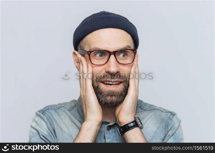 Amazed handsome middle aged man looks into distance in bewilderment, keeps hands on cheeks, wears glasses and hat, isolated over white background. People, emotions and unexpectedness concept