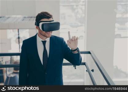 Amazed male office worker in suit being excited while trying out VR glasses, testing virtual reality for business, touching object with forefinger while exploring digital world, standing in office. Amazed male office worker in suit being excited while trying out VR glasses