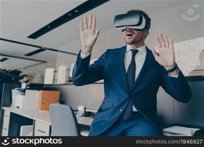 Amazed man in VR headset interacting with virtual objects or interface, sitting at edge of office table. Excited entrepreneur making 3D business presentation or meeting with virtual reality glasses. CEO man in VR headset interacting with virtual objects or interface while sitting in office
