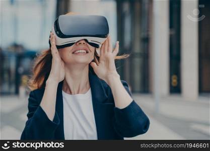 Amazed red haired female office worker being excited while trying out VR glasses, testing virtual reality for business, exploring digital world, standing alone against blurred urban background. Amazed red haired female office worker being excited while trying out VR glasses outdoors