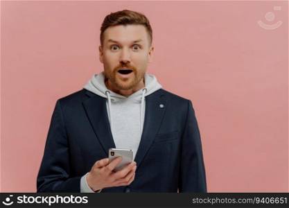 Amazed red-haired man in casual wear holds smartphone, shocked face, isolated on pastel pink background