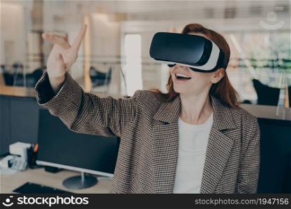 Amazed redhead female project manager reviewing virtual reality game, wearing VR glasses, touching object with her finger, excited businesswoman testing 3d goggles at work, interacting with cyberspace. Excited female project manager reviewing virtual reality game while working in office